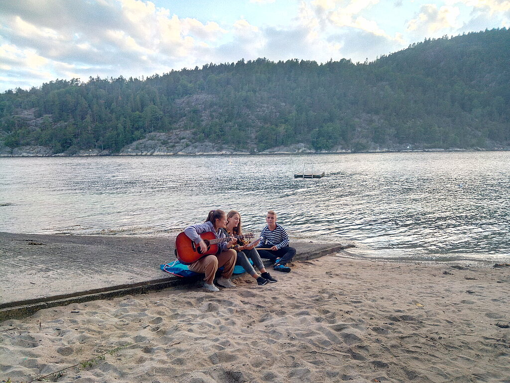 Drei Jugendliche sitzen auf einem Steg am Strand, eine spielt Gitarre.