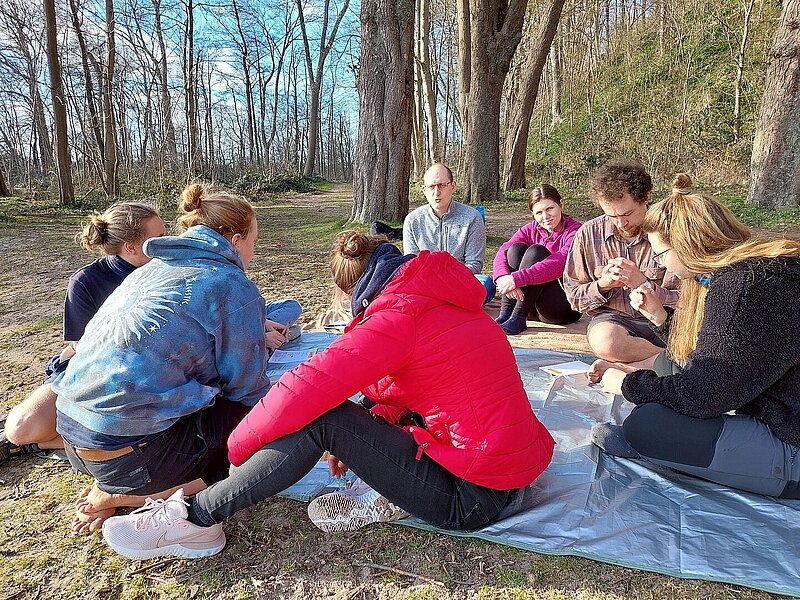 Eien Gruppe von jungen Menschen sitzt auf einer Decke in der Sonne, sie unterhalten sich miteinander.