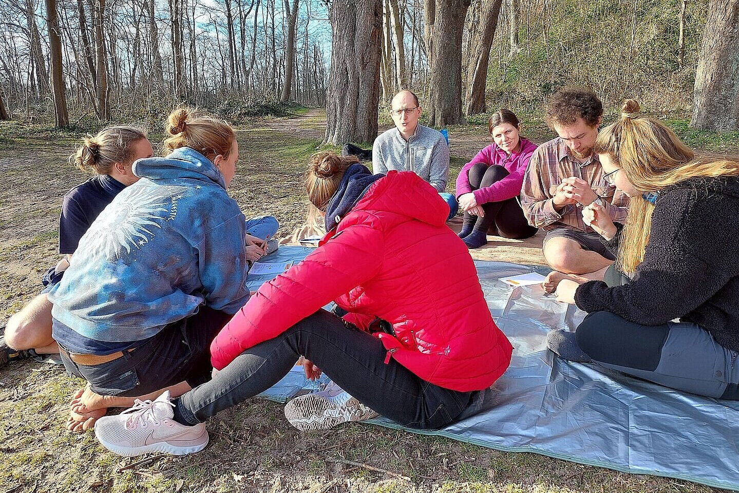 Eien Gruppe von jungen Menschen sitzt auf einer Decke in der Sonne, sie unterhalten sich miteinander.