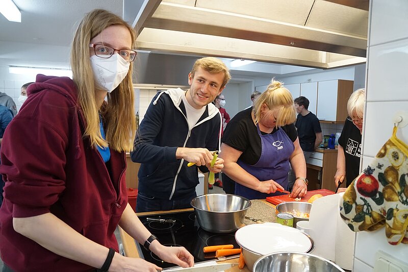 Einige Menschen schneiden Gemüse in der Küche, eine junge Frau schaut in die Kamera.