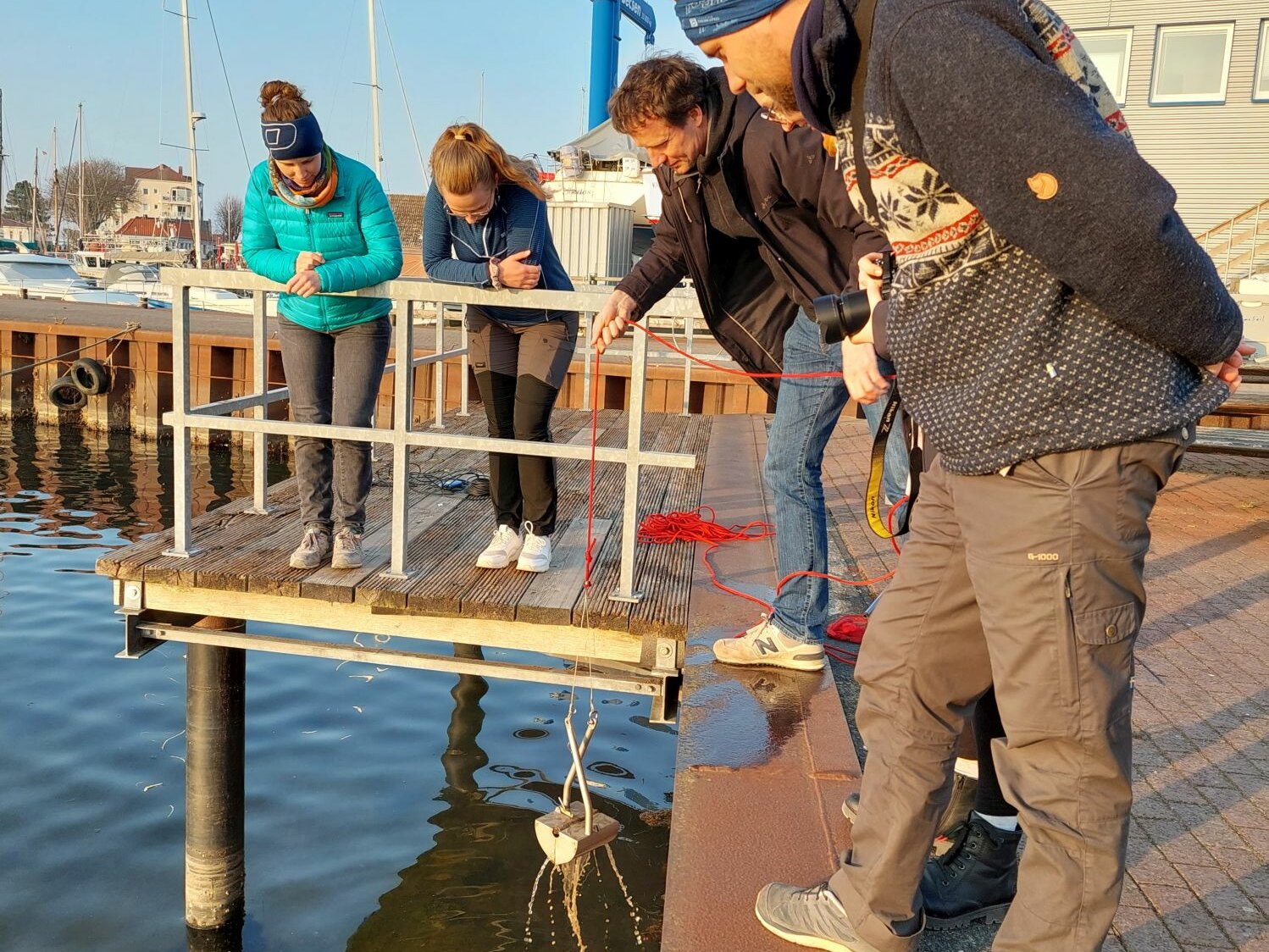 Fünf Menschen stehen an der Hafenkante und schauen vornüber gebeugt aufs Wasser. Einer von ihnen hebt ein Metallgerät aus dem Wasser.
