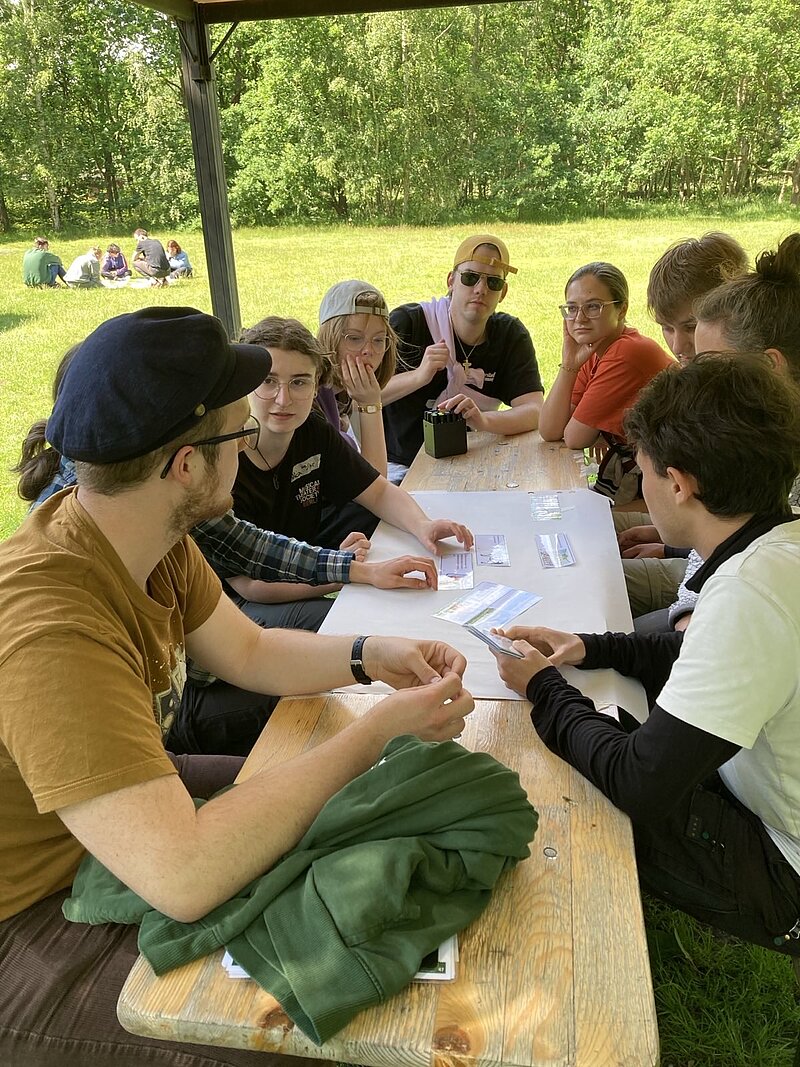 Einige junge Menschen sitzen um ein Flipchart und scheinen zu diskutieren und zu notieren.