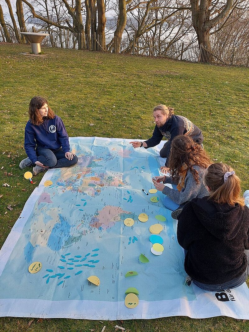 Vier junge Menschen sitzen auf einer großen Landkarte, die auf dem Rasen ausgebreitet ist. Einer von ihnen scheint etwas zu erklären.