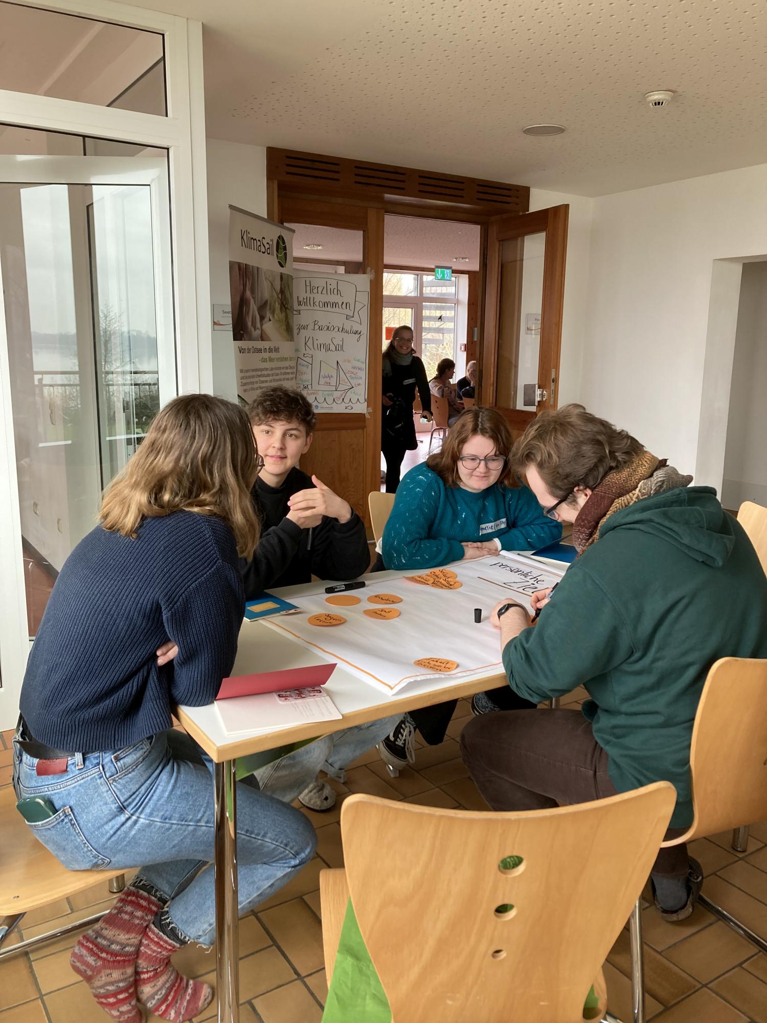 Vier junge Menschen sitzen um einen Tisch, auf dem eine Flipchart liegt. Sie scheinen sich zu unterhalten und Ideen zu sammeln.