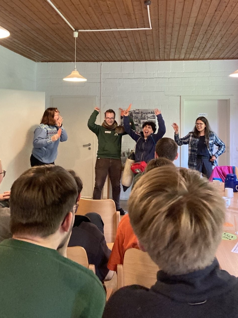 Vier junge Menschen stehen vor einer sitzenden Gruppe und nehmen theatralische Posen ein.