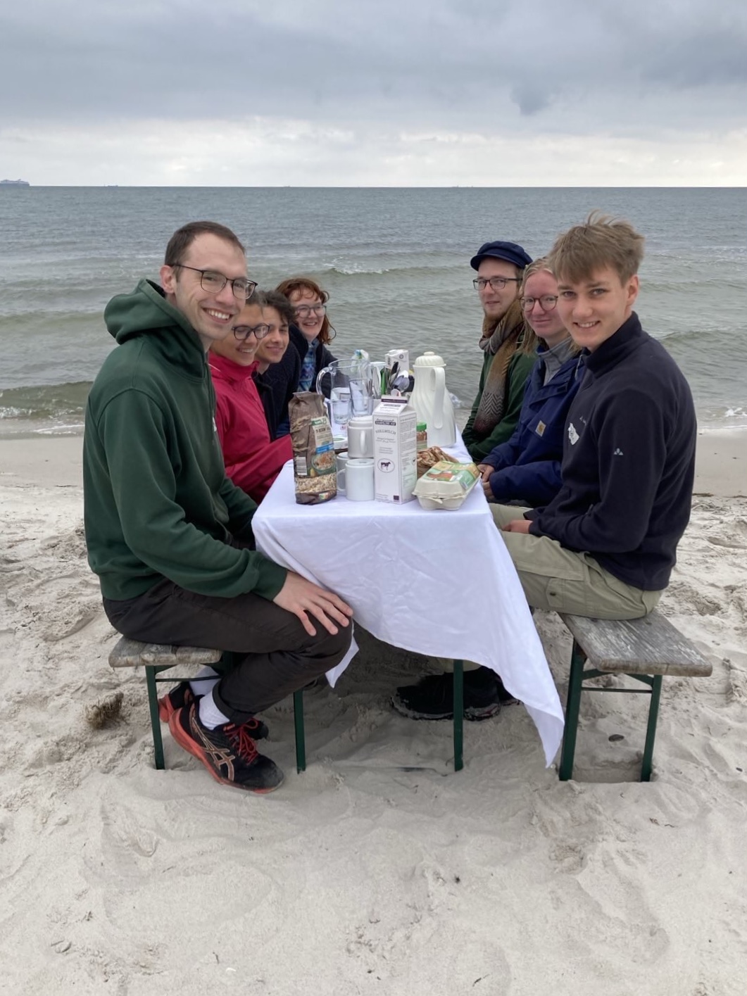 Einige junge Menschen sitzen an einem Tisch mit weißem Tischtuch am Strand, im Hintergrund Himmel und Ostsee.